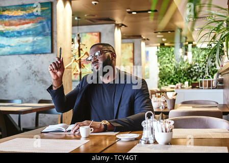 Portrait von stilvollen junge Afrikaner CEO in dunkel-blaue Jacke gehen durch etwas Schreibarbeit, sitzen im Cafe Tabelle, Notizen in Notebook, Whil Stockfoto