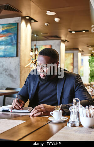 Portrait von stilvollen junge Afrikaner CEO in dunkel-blaue Jacke gehen durch etwas Schreibarbeit, sitzen im Cafe Tabelle mit Dokumenten und Tasse Kaffee Stockfoto