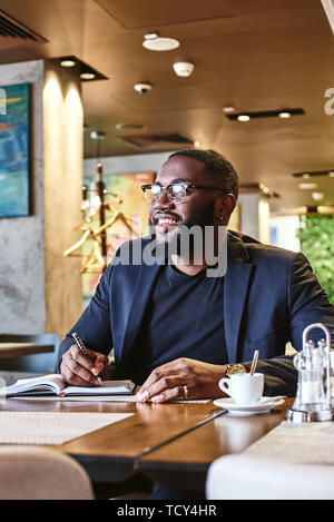 Portrait von stilvollen junge Afrikaner CEO in dunkel-blaue Jacke gehen durch etwas Schreibarbeit, sitzen im Cafe Tabelle mit Dokumenten und Tasse Kaffee Stockfoto