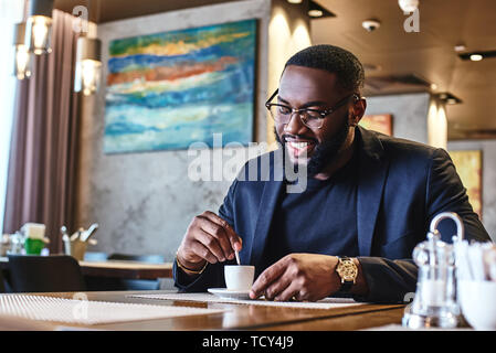 Stilvolle african american bärtigen Büroangestellter in Gläsern und dunkelblauen Anzug am Tisch sitzt. Er lächelt auf seine Tasse Espresso. Gemütliche i Stockfoto