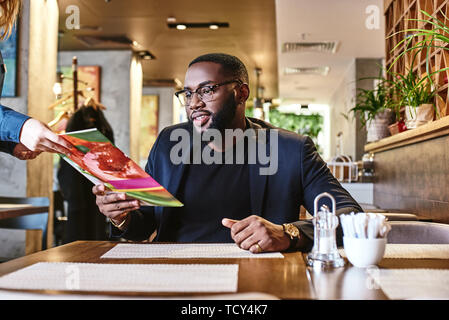 Stilvolle african american bärtigen Büroangestellter in dunkelblauen Anzug am Tisch sitzt. Er wählt, was er essen möchte und macht eine Bestellung während der Stockfoto