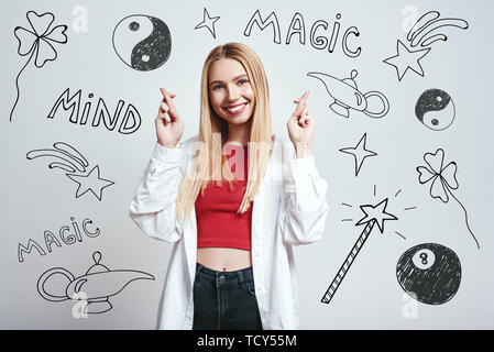 Ich weiß magic Trick. Lächelnde junge blonde Frau in weißem Hemd Holding die Daumen beim Stehen gegen grauen Hintergrund mit Hand gezeichnet Magie zu Stockfoto