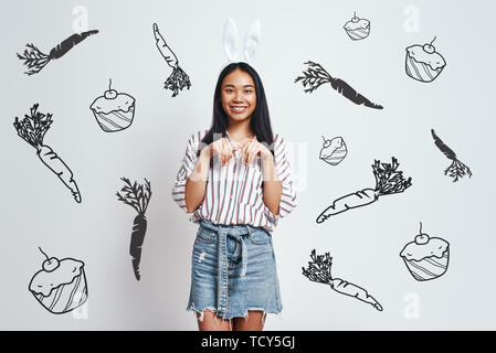 Funny Bunny. Nette junge asiatische Frau im bunny Ohren und Casual Wear lächelt, während gegen grauen Hintergrund stehend mit Hand gezeichnet Karotten und cupcak Stockfoto