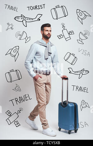 Travel Concept. Volle Länge des zuversichtlich und gut aussehender bärtiger Mann sein Gepäck beim Stehen gegen grauen Hintergrund mit Gekritzel auf der Sie Stockfoto