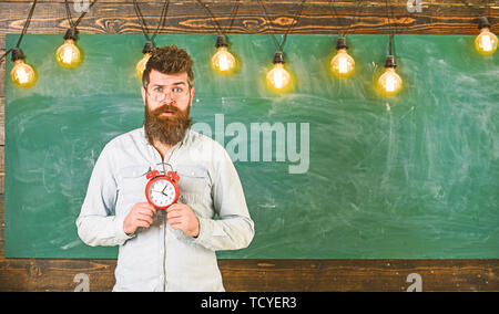 Lehrer in Brillen hält Wecker. Zeitplan und Regime Konzept. Bärtige hipster hält, schwarzen Brettes auf Hintergrund, kopieren. Mann mit Bart und Schnurrbart auf überrascht Gesicht im Klassenzimmer. Stockfoto