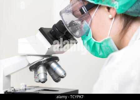 Junge Ärztin arbeiten mit dem Mikroskop im Labor, gegen Biohazard mit Maske, Hut und Brille. Stockfoto