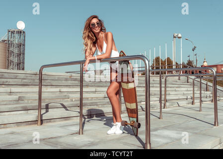 Glücklich lächelnd, skateboard Sommer in der Stadt. Ruhe sunbathes, posierend, weißen Körper Badeanzug, lange Haare gebräunte Abbildung. Freier Platz für Text. Emotionen Stockfoto