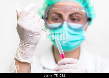 Junge attraktive Wissenschaftlerin in schützende Brillen, maske und handschuhe Fallen eine rote Flüssigkeit in das Reagenzglas mit einer Pipette im Labor konzentriert. Stockfoto