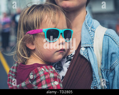 Eine kühle kleine kleinkind Sonnenbrille tragen wird in einer Schlinge von seiner Mutter durchgeführt Stockfoto