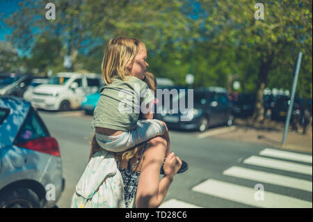 Ein kleines Kind reiten auf den Schultern seiner Mutter in einen Parkplatz auf einem sunnny Sommer Tag Stockfoto