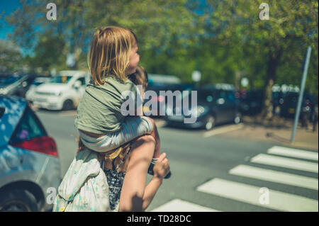 Ein kleines Kind reiten auf den Schultern seiner Mutter in einen Parkplatz auf einem sunnny Sommer Tag Stockfoto