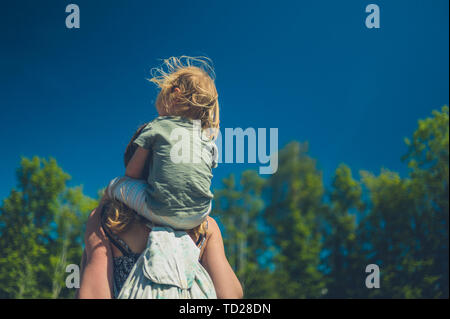 Ein kleines Kind reiten auf den Schultern seiner Mutter im Freien an einem sonnigen Sommertag Stockfoto