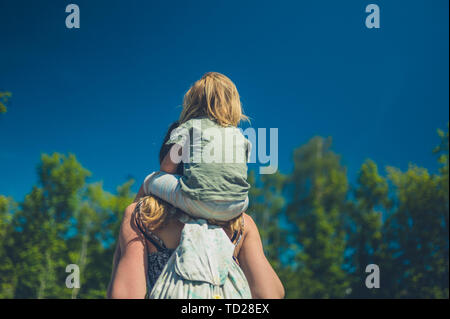 Ein kleines Kind reiten auf den Schultern seiner Mutter im Freien an einem sonnigen Sommertag Stockfoto