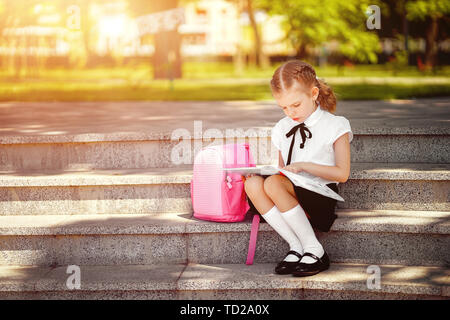 Schüler der Grundschule lesen Buch sitzen auf der Treppe. Mädchen mit Rucksack in der Nähe von Gebäude im Außenbereich. Beginn der Unterrichtsstunden. Erster Tag der Fall. Stockfoto