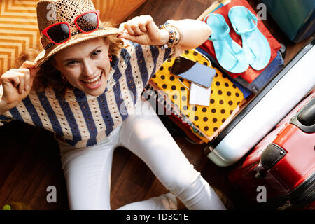 Obere Ansicht von lächelnden elegante Frau in weißen Hosen und gestreifte Bluse mit Abendmahl Hut und Sonnenbrille in der Nähe von Open travel Koffer im modernen Leben ro Stockfoto