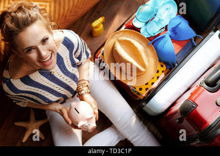 Obere Ansicht von lächelnden elegante Frau in weißen Hosen und gestreifte Bluse in der modernen Wohnzimmer im sonnigen Sommertag Holding piggy Bank in der Nähe von offenen Trave Stockfoto