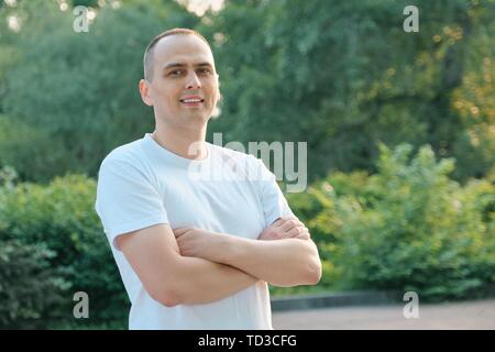 Outdoor Portrait der zuversichtlichen Lächeln Sport Mann mittleren Alters. Positive stattlicher Mann mit gekreuzten Armen im weißen T-Shirt in die Kamera schaut, backgro Stockfoto