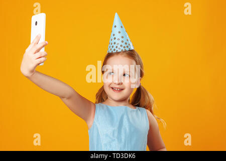 Fröhliches kleines Mädchen feiert Geburtstag. Das Kind hält das Telefon, nimmt eine selfie. Closeup Portrait auf gelben Hintergrund. Stockfoto