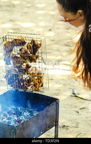 Frau mit langen dunklen Haaren halten Sie ein Raster von Grill mit Hühnerfleisch über glühende Holzkohle Stockfoto