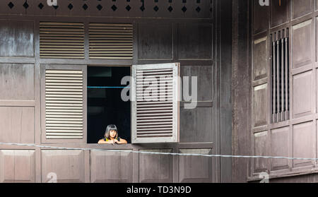 Makassar, Sulawesi, Indonesien - 28. Februar 2019: Kleines Mädchen sieht aus offenen Fenster, in dunklem Holz Wand gesetzt. Stockfoto