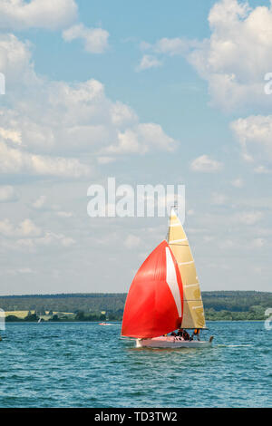 Eine kleine Freude Segelyacht mit Scarlet Sails geht an die Bucht auf einer verschlafenen Tag vor dem Hintergrund von einem dichten Wald auf dem gegenüberliegenden Ufer. Stockfoto