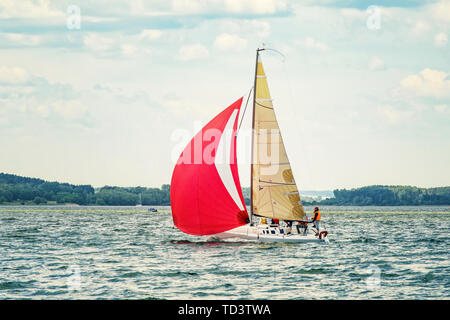 Eine kleine Freude Segelyacht mit Scarlet Sails geht an die Bucht auf einer verschlafenen Tag vor dem Hintergrund von einem dichten Wald auf dem gegenüberliegenden Ufer. Stockfoto