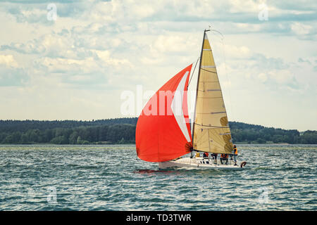 Eine kleine Freude Segelyacht mit Scarlet Sails geht an die Bucht auf einer verschlafenen Tag vor dem Hintergrund von einem dichten Wald auf dem gegenüberliegenden Ufer. Stockfoto