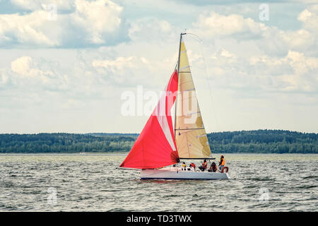 Eine kleine Freude Segelyacht mit Scarlet Sails geht an die Bucht auf einer verschlafenen Tag vor dem Hintergrund von einem dichten Wald auf dem gegenüberliegenden Ufer. Stockfoto