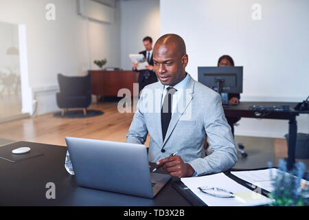 Junge afrikanische amerikanische Geschäftsmann an seinem Schreibtisch in einem Büro sitzen mit einem Laptop mit Kollegen im Hintergrund Stockfoto