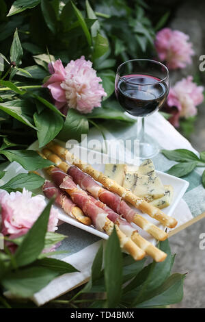 Apperitive mit einem Glas Rotwein, frischen Schinken, grissini Sticks und Käse in der Nähe von Pfingstrose blüht im Sommer. Stockfoto