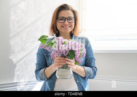 Portrait von schöne, reife Frau zu Hause mit Blumenstrauß aus lila Blüten. Hintergrund Licht wand Sonnenlicht im Fenster Stockfoto