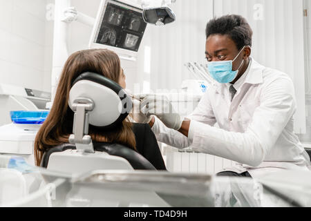 Arzt behandelt die Zähne. Junge afrikanische Mann Zahnarzt mit einem Patienten. Frau im Zahnarztstuhl an der Zahnklinik. Medizin, Gesundheit, Stomatologie Konzept Stockfoto
