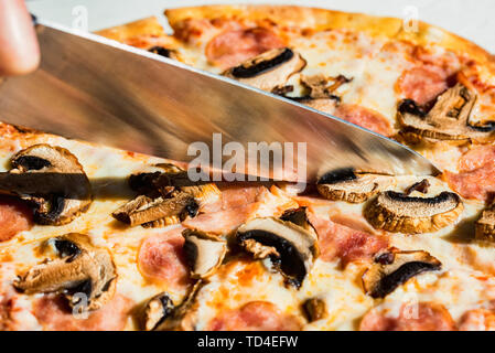 Close up Messer schneiden Pizza mit Salami, Pilzen und Käse aus der Nähe Stockfoto