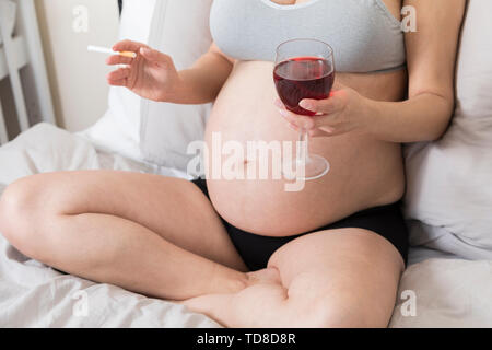 Junge Schwangere rauchen Zigaretten und trinken Alkohol, Frauen in der Schwangerschaft mit Tabak und Glas Wein, ungesunde Gewohnheiten und Missbrauch Stockfoto