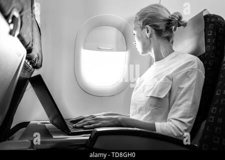 Attraktive kaukasische weiblichen Passagier auf der Suche durch die Ebene Fenster während der Arbeit an modernen Laptop Computer mit WLAN-Verbindung an Bord von Stockfoto
