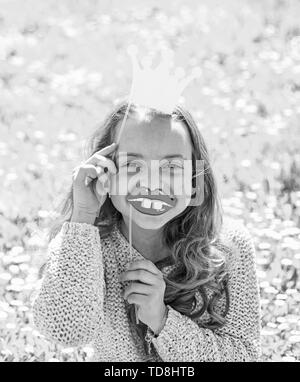 Mädchen sitzt auf Gras an grassplot, grünen Hintergrund. Kind posiert mit Pappe ein Lächeln auf den Lippen und Krone für Foto Session an der Wiese. Humor Königin Konzept. Mädchen auf fröhliche Gesicht verbringen Freizeitaktivitäten im Freien. Stockfoto