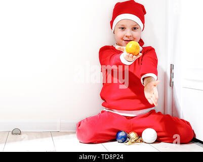 Ein kleiner Junge in der Form von Santa Claus lächelt, steht auf einem weißen Hintergrund, und hält eine Tangerine in seiner Hand. Stockfoto