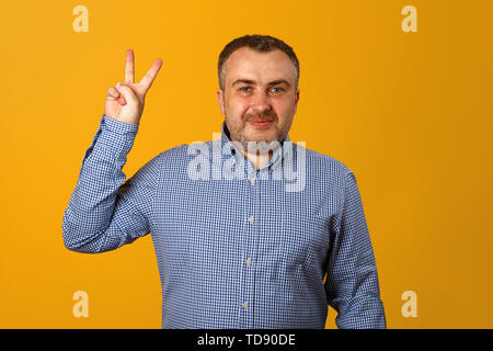 Ein junger Mann in einer Blue Plaid Shirt zeigt ein Sieg unterzeichnen und sieht in die Kamera. Gelber Hintergrund. Close-up. Stockfoto