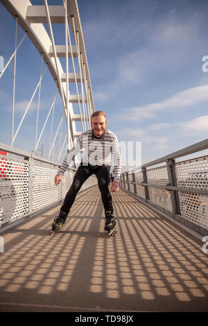 Ein junger Mann, Inline Skating im Freien auf einer Brücke. lächelnd in die Kamera schauen. Stockfoto