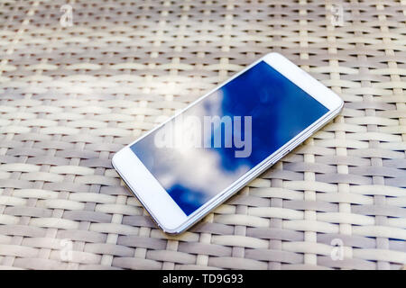 Reflexion der blauen Himmel mit Wolken im Telefon - Smartphone, das auf einem hellen Rattanmöbeln Tabelle vor dem Hintergrund liegt. Blur und Abstraktion Stockfoto