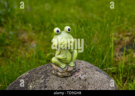 Ein Frosch auf einem Felsen Skulptur neben einem Teich in den Bäumen in einem Park mit dekorativer Frosch, Statue, Figur in Garten. Keramik Statue eines grünen Frosch. Stockfoto