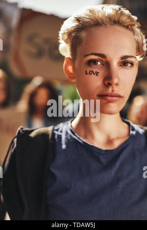 Nur Liebe. Der blonde junge Frau mit dem Wort Liebe auf ihrem Gesicht protestieren mit einer Gruppe von Aktivistinnen im Freien auf Straße geschrieben. Demonstration Stockfoto