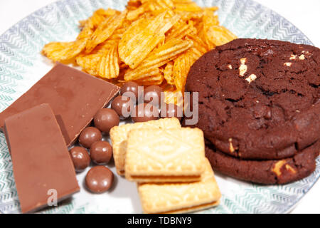Platte aus Triple-Chocolate-Plätzchen, mit Schokolade überzogene Wabenkugeln, Crinkle Cut Crisps, Schokoriegel auf einer weißen Platte mit grell gestreifter Bekantung Stockfoto