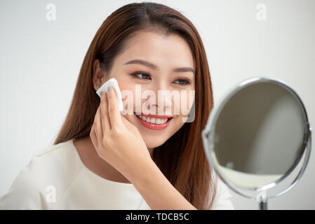 Nehmen Sie Ihr Make-up. Schöne fröhliche junge Frau mit Baumwolle Scheibe und Blick auf ihr Spiegelbild im Spiegel mit Lächeln und das Kleid sitzt Stockfoto