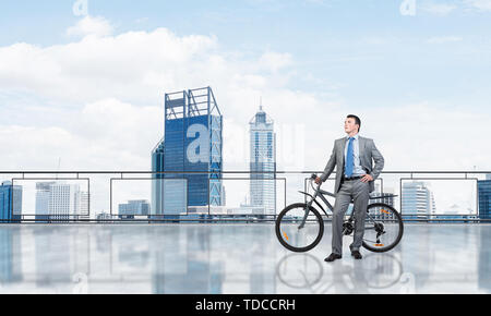 Junger Mann mit Anzug stehen auf Balkon mit dem Fahrrad. Geschäftsmann mit dem Fahrrad auf dem Hintergrund des blauen Himmels über Megalopolis. Männliche Radfahrer halten Stockfoto