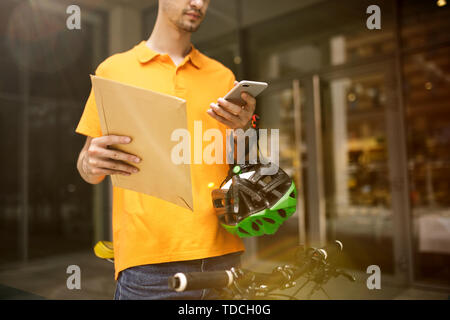 Junger Mann im gelben Trikot, Paket mit Gadgets um an der Hauptstraße der Stadt zu verfolgen. Kurier online App für den Empfang der Zahlung und Versand Adresse. Moderne Technologien. Stockfoto