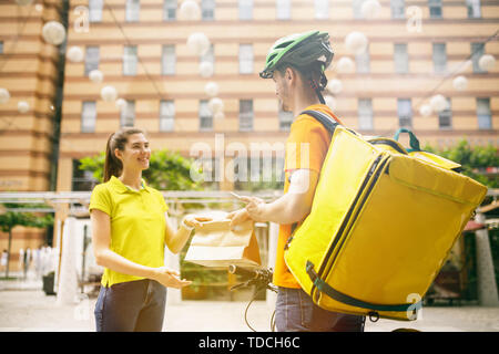 Junger Mann im gelben Trikot, Paket mit Gadgets um an der Hauptstraße der Stadt zu verfolgen. Kurier online App für den Empfang der Zahlung, die Adresse und die Empfänger. Moderne Technologien. Stockfoto