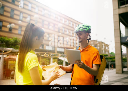 Junger Mann im gelben Trikot, Paket mit Gadgets um an der Hauptstraße der Stadt zu verfolgen. Kurier online App für den Empfang der Zahlung, die Adresse und die Empfänger. Moderne Technologien. Stockfoto