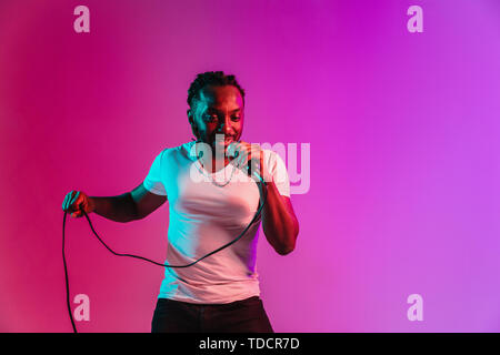 Junge afrikanische-amerikanischen Jazzmusiker mit Mikrofon singen ein Lied auf lila studio Hintergrund in trendigen Neon Licht. Konzept aus Musik, Hobby, inspirness. Bunte Porträt der freudigen attraktive Künstler. Stockfoto