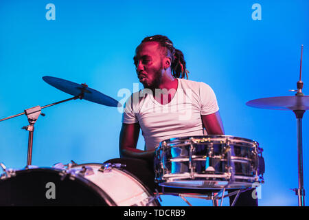 Junge afrikanische-amerikanischen Jazz Musiker oder Drummer Schlagzeug spielen auf Blau studio Hintergrund in trendigen Neonlicht. Konzept aus Musik, Hobby, inspirness. Bunte Porträt der freudigen attraktive Künstler. Stockfoto